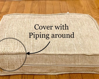 May include: Beige cushion cover with piping around the edges. The cover has a zipper closure.