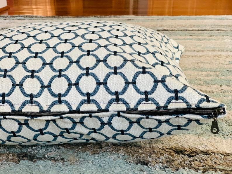 K&ouml;nnte beinhalten: Ein wei&szlig;-blaues, gemustertes Bodenkissen mit Rei&szlig;verschluss. Das Kissen ist aus weichem, bequemem Stoff und hat ein geometrisches Muster.