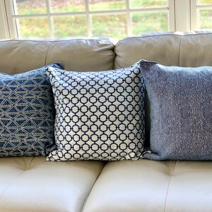 K&ouml;nnte beinhalten: Drei dekorative Kissen mit blauen und wei&szlig;en geometrischen Mustern. Die Kissen liegen auf einem wei&szlig;en Ledersofa.