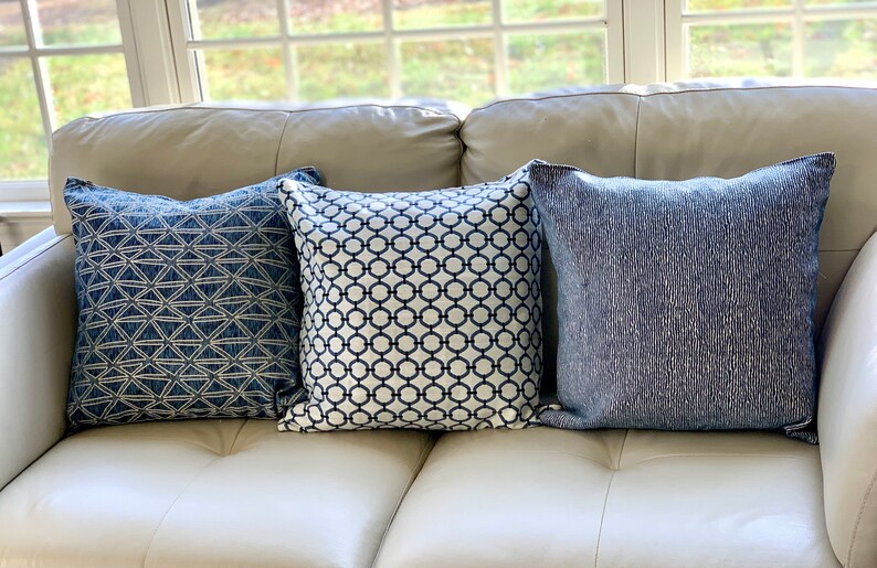 K&ouml;nnte beinhalten: Drei dekorative Kissen mit blauen und wei&szlig;en geometrischen Mustern. Die Kissen liegen auf einem wei&szlig;en Ledersofa.