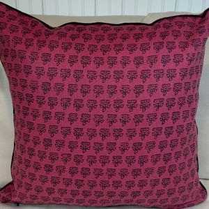 May include: A square decorative pillow with a magenta background and a repeating black floral pattern. The pillow has a black trim around the edges. The pillow is likely used for home decor.