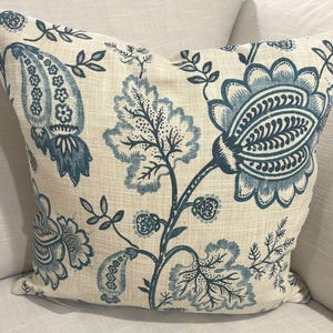 Puede incluir: Una almohada de color crema con un patrón floral azul. La almohada presenta una gran flor azul con detalles intrincados y flores y hojas azules más pequeñas en todo el diseño.