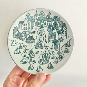 Könnte beinhalten: Ein runder, weißer Keramikteller mit einer teal-farbenen Illustration einer Küstenstadt. Das Design umfasst Häuser, Bäume, Menschen und Segelschiffe. Der Teller hat einen dünnen teal-farbenen Rand.