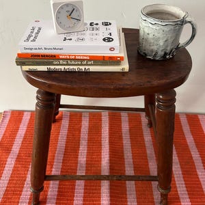 May include: A small, round, dark wooden stool with a stack of books, a white clock, and a ceramic mug. The books include "Design as Art," "Ways of Seeing," and "Modern Artists On Art." The stool sits on a striped orange and pink rug.