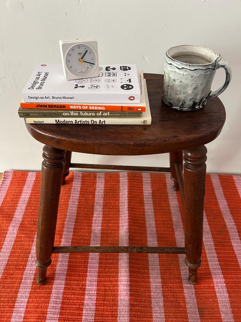 May include: A small, round, dark wooden stool with a stack of books, a white clock, and a ceramic mug. The books include "Design as Art," "Ways of Seeing," and "Modern Artists On Art." The stool sits on a striped orange and pink rug.