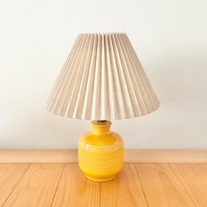 Könnte beinhalten: Eine kleine Tischlampe aus gelber Keramik mit beigefarbenem, plissiertem Lampenschirm. Der Lampenfuß ist leuchtend gelb mit horizontalen Streifen. Der Lampenschirm ist hellbeige und konisch geformt.