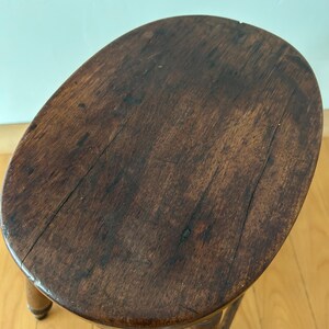 May include: A small, antique wooden stool with a dark brown, oval-shaped seat. The seat shows visible wood grain and wear. The stool has three legs and a supporting stretcher, all made of the same dark wood. The stool is a vintage piece.