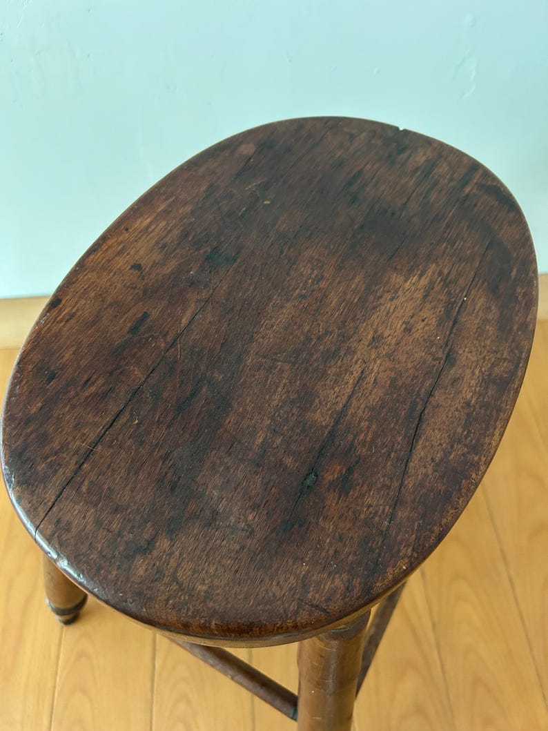 May include: A small, antique wooden stool with a dark brown, oval-shaped seat. The seat shows visible wood grain and wear. The stool has three legs and a supporting stretcher, all made of the same dark wood. The stool is a vintage piece.
