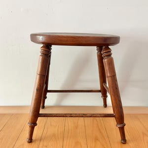 May include: A vintage wooden stool with a round seat and four turned legs. The stool is made of dark-stained wood, showcasing the natural grain. A horizontal stretcher connects the legs for added stability. The stool is a classic piece of furniture.