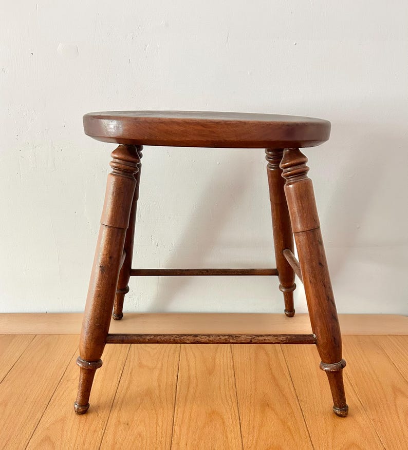 May include: A vintage wooden stool with a round seat and four turned legs. The stool is made of dark-stained wood, showcasing the natural grain. A horizontal stretcher connects the legs for added stability. The stool is a classic piece of furniture.