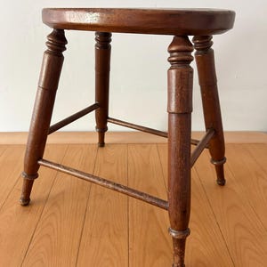 May include: A vintage wooden stool with a round seat and four turned legs. The stool is stained a warm brown color, and the legs are connected by horizontal supports. The design is simple and classic, suitable for various interior styles.