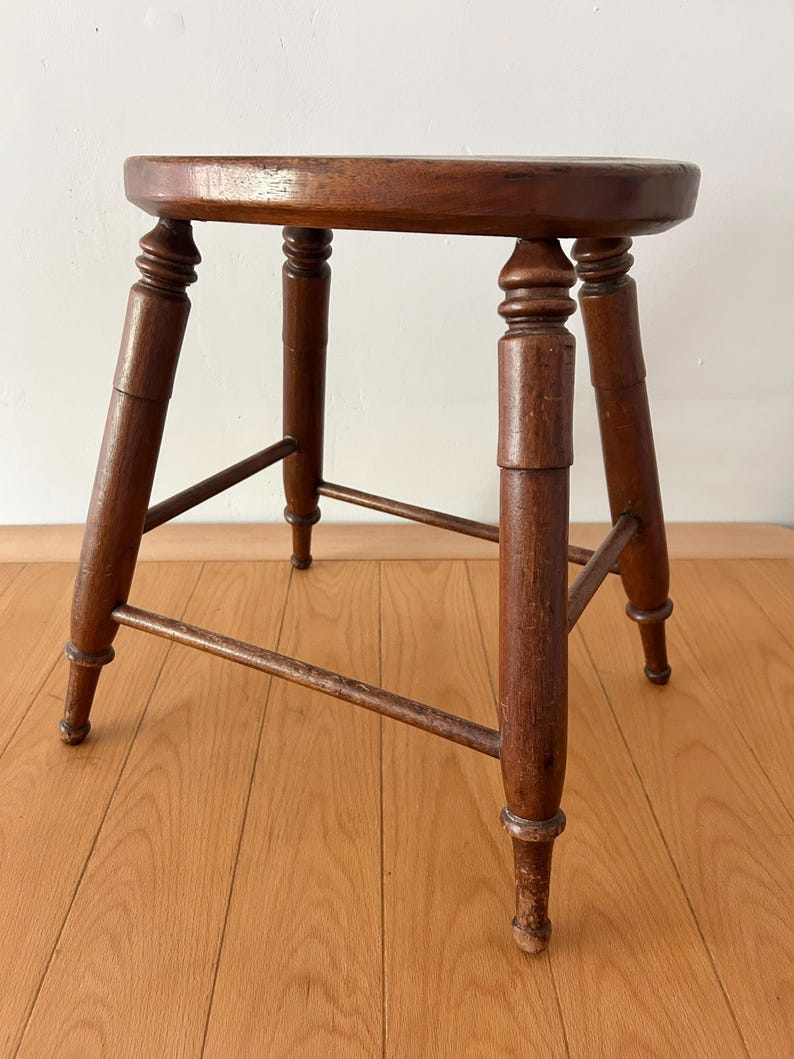 May include: A vintage wooden stool with a round seat and four turned legs. The stool is stained a warm brown color, and the legs are connected by horizontal supports. The design is simple and classic, suitable for various interior styles.