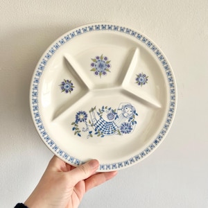 Może przedstawiać: Biały, ceramiczny talerz podzielony na sekcje z niebieskim i zielonym wzorem kwiatowym oraz stylizowaną ilustracją kobiety. Talerz ma trzy sekcje.