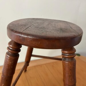 May include: A vintage wooden stool with a round seat and four turned legs. The stool is stained a dark brown color, showing signs of wear and age. The seat has visible cracks, adding to its rustic charm. The legs are connected by a lower stretcher.