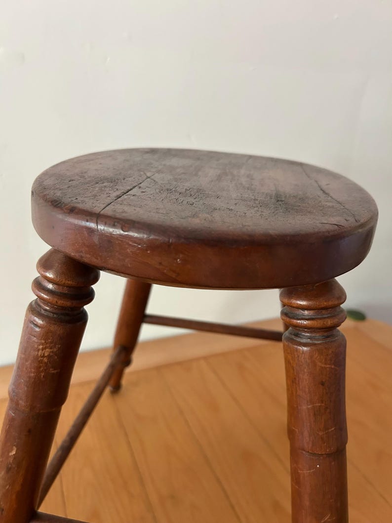 May include: A vintage wooden stool with a round seat and four turned legs. The stool is stained a dark brown color, showing signs of wear and age. The seat has visible cracks, adding to its rustic charm. The legs are connected by a lower stretcher.