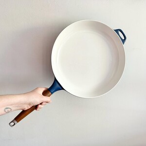 May include: A white enamelled cast iron skillet with a blue rim and a wooden handle. The skillet is being held by a hand.
