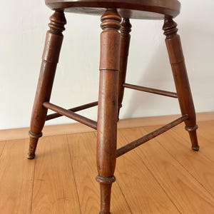 May include: A small, antique wooden stool with a round seat and four turned legs. The stool is made of dark brown wood and has a classic design. The legs are connected by horizontal supports for added stability. The stool is approximately 18 inches tall.