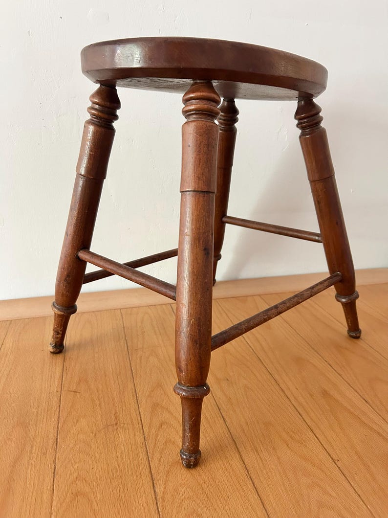 May include: A small, antique wooden stool with a round seat and four turned legs. The stool is made of dark brown wood and has a classic design. The legs are connected by horizontal supports for added stability. The stool is approximately 18 inches tall.