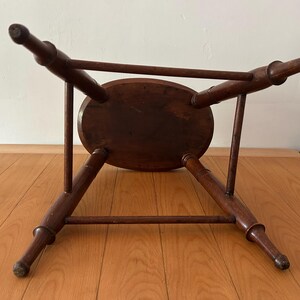 May include: A dark brown, antique wooden stool with an oval seat and four splayed legs. The legs are connected by a horizontal stretcher. The stool is viewed from underneath, showcasing its construction and design.