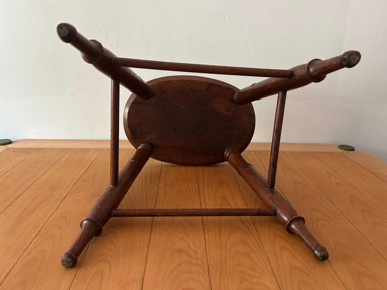May include: A dark brown, antique wooden stool with an oval seat and four splayed legs. The legs are connected by a horizontal stretcher. The stool is viewed from underneath, showcasing its construction and design.