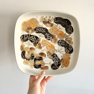 Può includere: Un piatto da portata quadrato in ceramica bianca con un motivo floreale marrone e oro. Il piatto ha un bordo rialzato e una superficie liscia.