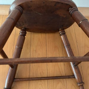 May include: A vintage wooden stool with a round seat and four turned legs. The stool is made of dark-stained wood, showing signs of age and wear. The legs are connected by a lower stretcher for added stability. The stool is positioned on a wooden floor.
