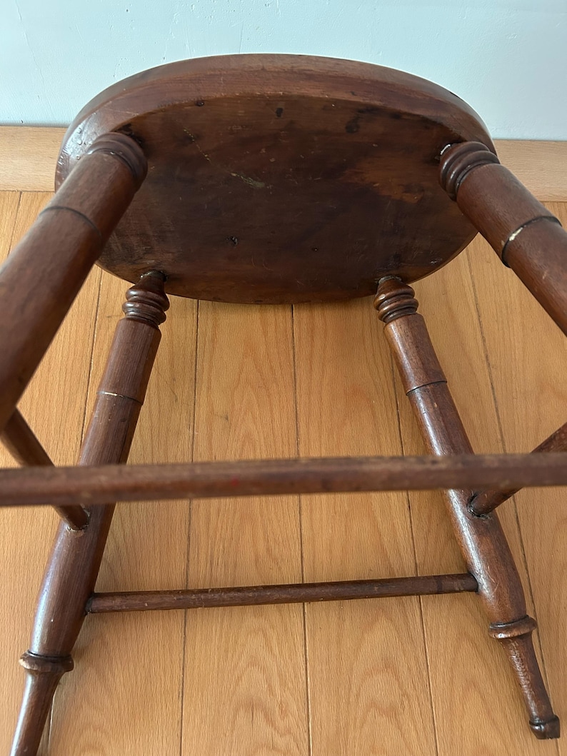 May include: A vintage wooden stool with a round seat and four turned legs. The stool is made of dark-stained wood, showing signs of age and wear. The legs are connected by a lower stretcher for added stability. The stool is positioned on a wooden floor.
