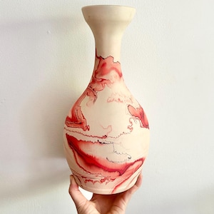 Peut inclure: Un vase en céramique de couleur crème avec un dessus évasé et un corps arrondi. Le vase est décoré de motifs abstraits rouges et noirs fluides. Le vase est tenu par une main sur un fond blanc.