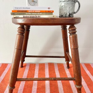May include: A wooden stool with a round top, holding a stack of books and a mug. The books include "Design as Art," "Ways of Seeing," and "Modern Artists On Art." A small white clock sits atop the books. The stool is on a striped orange and pink rug.