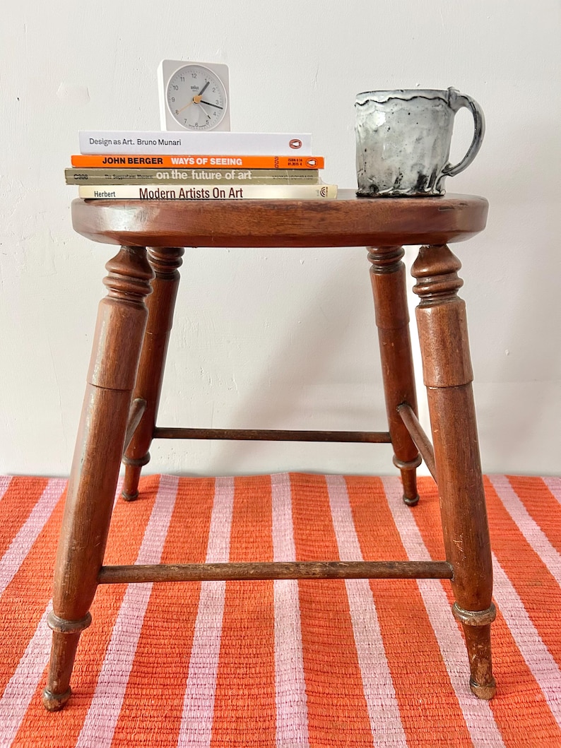 May include: A wooden stool with a round top, holding a stack of books and a mug. The books include "Design as Art," "Ways of Seeing," and "Modern Artists On Art." A small white clock sits atop the books. The stool is on a striped orange and pink rug.