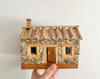 May include: A miniature stone house with a wooden door and windows. The roof is made of textured, light brown tiles, and a small chimney is visible. The house is held in a hand against a white background.