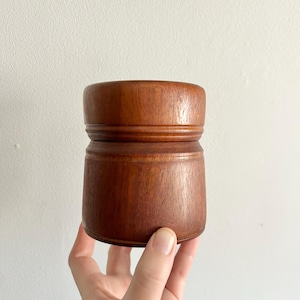 Può includere: Un piccolo contenitore rotondo in legno con una finitura marrone scuro. Il contenitore ha un design semplice con una base leggermente svasata e una superficie liscia e lucida.
