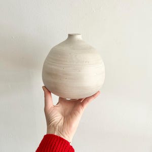 May include: A white ceramic vase with a round body and a narrow opening. The vase has a speckled finish and is being held in a hand.