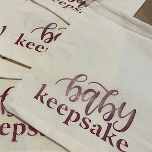 May include: A stack of white cotton drawstring bags with the text "baby keepsake" printed in maroon on the front.