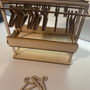 May include: A wooden dollhouse-sized clothing rack with a shelf and a row of wooden hangers. There are two additional wooden hangers on the white surface below the rack.