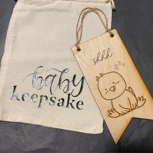 Puede incluir: Un adorno de madera para bebé con un lindo pollito de dibujos animados y el texto "shhhh zzz". El adorno está sujeto a una cuerda y cuelga de una bolsa de lona blanca con el texto "baby keepsake" impreso en ella.