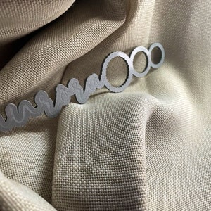 May include: A silver metal key chain with a wavy design and three circular rings. The keychain is lying on a beige fabric surface.