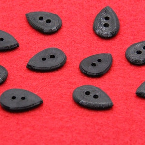 May include: A collection of ten black, teardrop-shaped buttons with two holes each, arranged on a vibrant red background. The buttons have a textured appearance, suggesting a 3D-printed design, and are suitable for sewing or crafting projects.