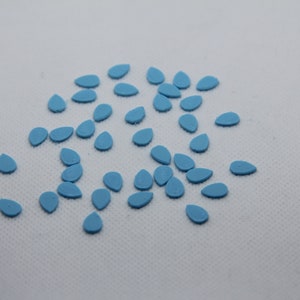 Puede incluir: Una dispersión de pequeños adornos en forma de lágrima de color azul claro. Estos elementos decorativos están dispersos sobre una superficie blanca. Las lágrimas son de tamaño y forma uniformes, con un acabado liso y mate.