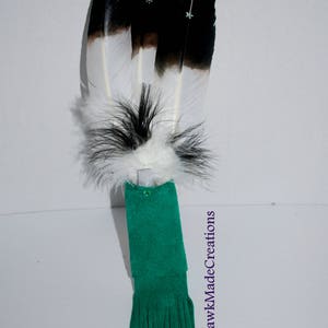 May include: A green suede feather fan with black and white feathers and a fringe. The fan is decorated with small, iridescent beads. "MohawkMadeCreations" is written on the right side of the image.