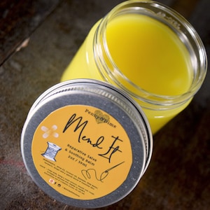 May include: A small glass jar with a silver lid containing a yellow salve. The label on the jar reads "Penny & Pine Mend It Reparative Salve & Soothing Balm 2 oz / 59ml".