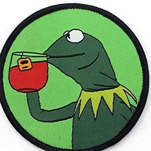 Może przedstawiać: Okrągła, haftowana naszywka przedstawiająca Kermita Żabę pijącego z kieliszka z czerwonym płynem. Naszywka ma zielone tło i czarną obwódkę. Kermit jest zielony z żółtym kołnierzem.