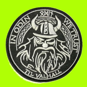 May include: A round, black and white embroidered patch featuring a Viking warrior with a horned helmet and long beard. The text "IN ODIN WE TRUST TIL VALHALL" is embroidered around the image. The patch is set against a bright green background.