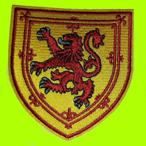 May include: Embroidered patch featuring a red lion rampant on a yellow shield. The shield has a red border and decorative elements. The lion has blue accents on its claws and tongue. The patch is likely for clothing or accessories.