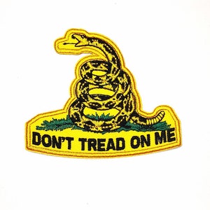 Peut inclure: Écusson brodé représentant un serpent à sonnettes enroulé, jaune et noir, sur de l'herbe verte, avec un bord jaune. Le texte "DON'T TREAD ON ME" est affiché en lettres majuscules noires sur une bannière jaune.