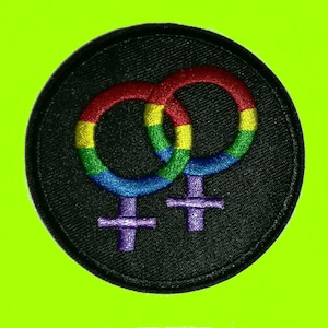 Puede incluir: Un parche redondo y negro bordado con dos símbolos femeninos entrelazados de color arcoíris. Los símbolos son rojos, naranjas, amarillos, verdes y azules. El parche tiene un borde cosido y está sobre un fondo verde brillante.