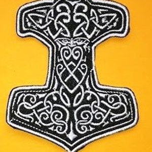 Może przedstawiać: Haftowana naszywka przedstawiająca stylizowany, czarno-biały młot Thora. Młot ma skomplikowane wzory węzłów i centralny motyw twarzy na żółtym tle. Przeznaczona do odzieży lub akcesoriów.