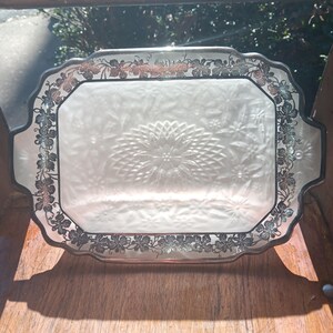 Puede incluir: Bandeja rectangular con superficie de vidrio esmerilado y borde decorativo plateado. El borde presenta un diseño de vid. La bandeja tiene asas a cada lado y un motivo floral central.