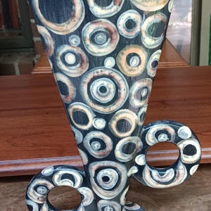 May include: A tall, flared ceramic vase featuring a dark charcoal background covered in a dense pattern of concentric circles in shades of cream, tan, and muted orange. The vase has two circular, open-loop handles on the sides and a round base.