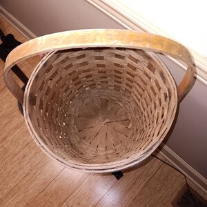 May include: A round, woven basket with a wooden handle. The basket is made of light brown woven material and has a circular shape. The handle is a light tan color.